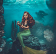 A beautiful mermaid at the bottom of the sea sits on a stone and holds a shell with a pearl