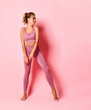 Beautiful young female athlete wearing pink stands leaning on one leg looking aside with laugh. Full length portrait isolated on rose, copy space