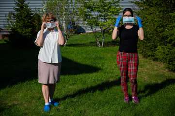 Mom and daughter put on medical masks observing the social distance. Two women stand in the park at a great distance from each other. Against the spread of the virus.