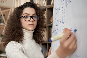 Young serious female latin math school teacher wearing glasses holding writing equation on whiteboard in classroom. Hispanic university college tutor, graduate student learning, teaching during class.