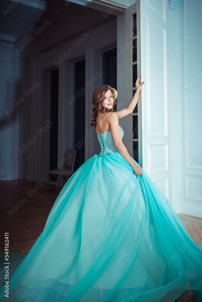 The beautiful young girl the princess in a long magnificent turquoise dress stands in a light castle. Fairytale fashion photo session Cinderella. Beautiful room. Princess runs at the door Stock Photo