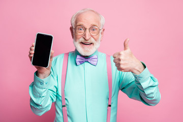 Portrait of amazed excited old man hold new smartphone show thumb up sign recommend suggest select wear teal turquoise shirt purple bowtie isolated over pink pastel color background