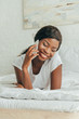 © LIGHTFIELD STUDIOS - surface level of cheerful african american girl talking on smartphone while lying on white bedding