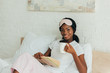 © LIGHTFIELD STUDIOS - happy african american girl with sleep mask on forehead sitting in bed, holding book and cup of coffee