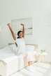 © LIGHTFIELD STUDIOS - happy african american girl in pajamas stretching with raised hands while sitting on bed