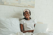 © LIGHTFIELD STUDIOS - happy african american freelancer with sleep mask on forehead using laptop in bed and smiling at camera