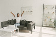 © LIGHTFIELD STUDIOS - happy african american freelancer stretching while sitting on sofa near table with laptop in spacious living room