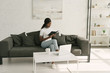 © LIGHTFIELD STUDIOS - attentive african american freelancer reading notebook while sitting on sofa near table with laptop