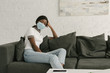 © LIGHTFIELD STUDIOS - thoughtful african american girl in medical mask sitting on grey sofa at home