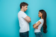 © LIGHTFIELD STUDIOS - side view of offended interracial couple standing with crossed arms and looking at each other on blue background