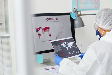 Wall Mural - Rear view of doctor in protective clothing sitting at her workplace in front of computer and looking at covid statistics