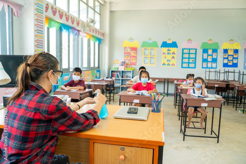 Fotomural Teacher wearing protective mask to Protect Against Covid-19,Group of school kids with teacher sitting in classroom online and raising hands,Elementary school,Learning and people concept