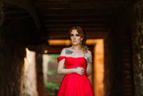 Fototapeta Londyn - Attractive redhead tattooed woman in red dress and diadema posing on blurred medieval castle background