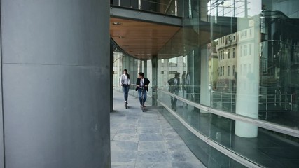 Wall Mural - Two smiling business people driving electric scooter in front of modern business building going on work.