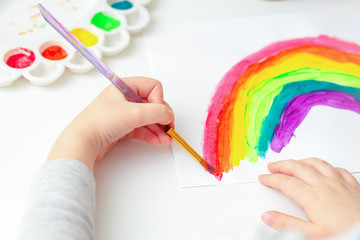 Sticker - Top view of hands of child drawing a rainbow with brushes and paints on white paper.