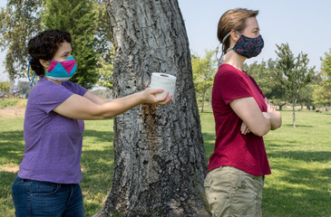 a Caucasian woman wearing a face mask offering a roll of toilet paper to her angry partner