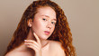 © Shauerman - beautiful girl with red long curly hair and brown eyes without makeup looks at the camera. Studio shot on a beige background, close-up. The concept of natural beauty, young skin care
