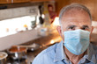 Ritratto di uomo anziano con mascherina protettiva e camicia in jeans da dentro la sua cucina