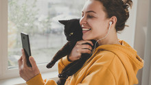 Cheerful Young Woman Wearing Headphones Holds Black Pet Cat Using Smartphone For Video Call, Gesturing Hi To Friend Or Parent. Happy Smiling Hipster Girl Making Facetime Zoom Video Calling