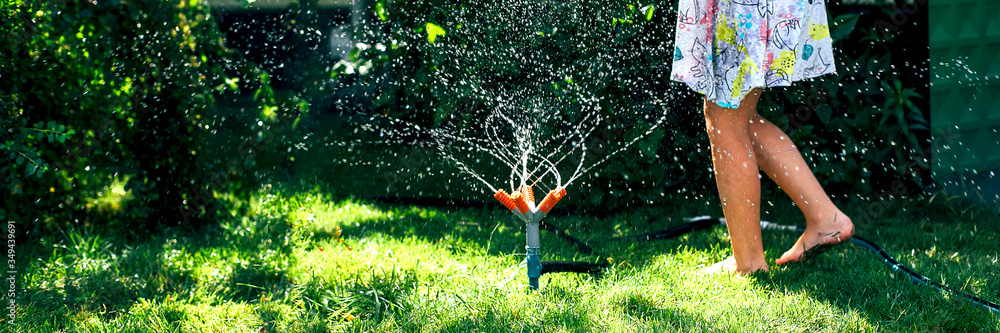 Child playing with garden sprinkler. Happy Girl in dress running and jumping. Kids gardening. Summer outdoor water fun. Children play with gardening hose watering flowers. - obrazy, fototapety, plakaty 
