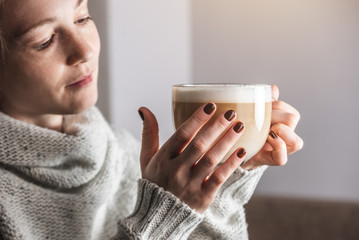 A young beautiful woman in a light warm sweater is sitting in a cozy cafe with a cup of hot delicious and aromatic latte. Concept of breakfast or break for a cup of coffee