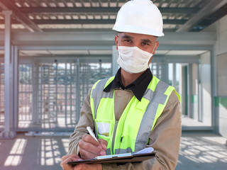 Construction worker in white hardhat and protective mask stands with clipboard opposite checkpoint with turnstile