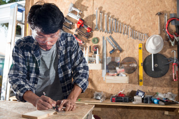 The carpenter is drawing lines on the wood that is placed on the wooden table. And there are many mechanics hanging on the wall