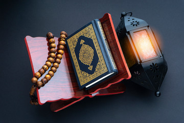 Poster - Holy Al Quran with written arabic calligraphy meaning of Al Quran, lantern lamp and rosary beads or tasbih on black background.