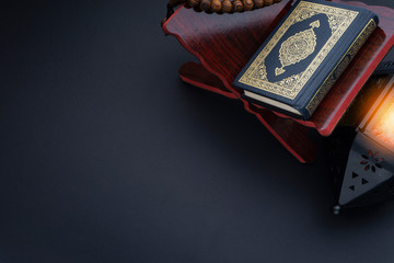 Holy Al Quran with written arabic calligraphy meaning of Al Quran, lantern lamp and rosary beads or tasbih on black background.