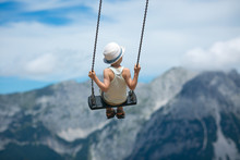 A Boy In A Summer Hat Flies On The Swings On A Background Of Rocky Mountains