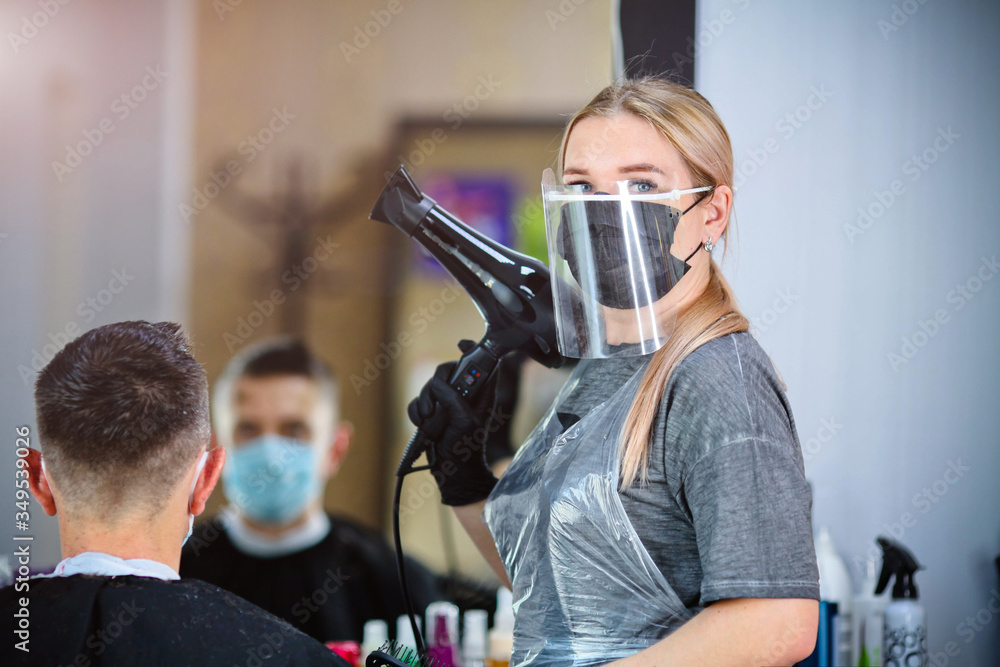 A hairdresser with security measures for Covid-19 holding a hairdryer and looking at the camera in a medicine mask, social distance, cutting hair with a medical mask, eye mask and  gloves - obrazy, fototapety, plakaty 