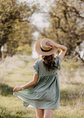 Young woman standing on a tree tunnel background, outdoors in a park, wearing a vintage light green dress and a straw hat.