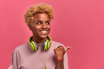 Wall Mural - Close-up portrait of cheerful African American student pointing with thumb to the left on pink background in studio. Concept of discounts and special offers in a store for youth. Place for text