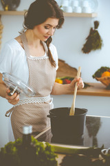 Wall Mural - Young brunette woman cooking soup in kitchen. Housewife holding wooden spoon in her hand. Food and health concept