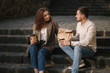 © Aleksandr - Young couple takaaway food and sit outdoors on stairs in city. Stylish couple drink coffe and wait friends