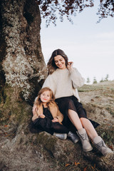 Wall Mural - Stylish young family in the autumn mountains. Mom and daughter are sitting under a large old tree and hugging against the background of the forest and mountain peaks at sunset.