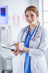 Wall Mural - Young woman doctor is standing with board with clipboard smiling in hospital office