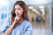 © whyframeshot - Beautiful Asian female patient hospital uniform looking at camera making surprise and quiet hand sign forefinger touch on lips gossip concept with clinic hospital blur background