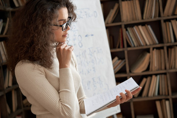 Young female latin math school teacher wearing glasses holding notebook thinking on task in classroom. Hispanic university college tutor, graduate student learning or teaching during class in campus.