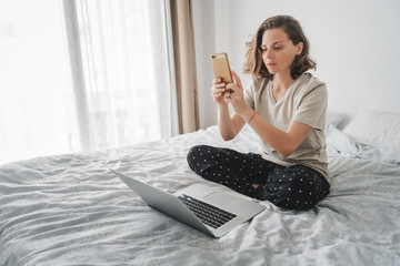 Wall Mural - Pretty young curly woman spends time with a smartphone and a laptop on the bed in the bedroom, 