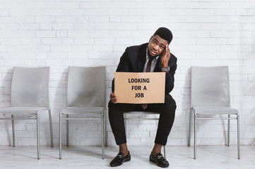 Wall Mural - African American guy with LOOKING FOR JOB sign sitting in company foyer, waiting for work interview. Free space