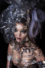 Close Up Portrait Of Young Graceful Nude Girl With Professional Black And Gold Hieroglyph Body Painting. Sexy Beautiful Expressive Woman With Garbage And Foil Transparent Dress And Lush Hairstyle