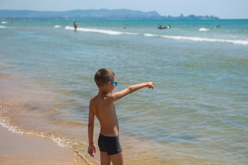 Canvas Print - A child in sunglasses and swimming trunks looks at the ocean. A young boy points at the sea.