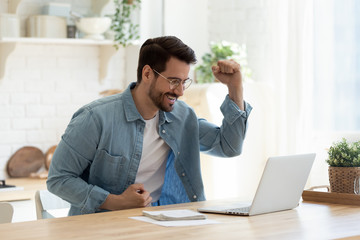 Excited millennial man in glasses sit at table in kitchen feel euphoric win online lottery on laptop, overjoyed young male in spectacles look at computer screen triumph get good news on email