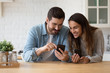 © fizkes - Smiling millennial couple sit at table in kitchen have fun using modern smartphone devices together, happy young husband and wife laugh relax at home browsing application on cellphone gadgets