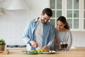 Wall Mural - Happy millennial husband and wife stand at counter cooking dinner salad together, smiling young Caucasian couple have fun preparing food in home kitchen enjoy family weekend or celebration