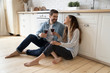 © fizkes - Happy millennial couple sit relax on wooden floor in kitchen drinking red wine talking chatting, smiling young husband and wife rest at home enjoy romantic date on family weekend together
