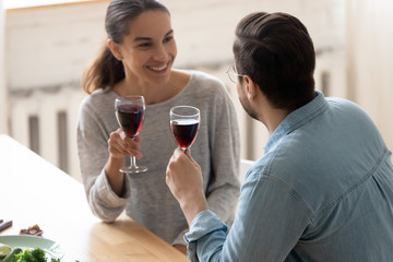 Wall Mural - Smiling young husband and wife sit in modern kitchen drink red wine enjoy family leisure domestic weekend together, happy millennial couple celebrate wedding anniversary or romantic date at home