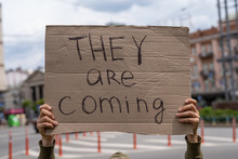 Suspicious And Warning Inscription On Sign. They Are Coming. Hands Holding Banner Outside On City Streets. Social Protest Motivation Concept. Guy With Sign Imitation. Aliens Guests Waiting Invitation