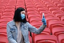 A Cheerleader Girl In A Medical Mask And Rubber Gloves Takes A Selfie Or Photographs On A Smartphone Alone In An Empty Stadium With Red Seats. Cancellation Of Sporting Events During The Coronavirus.